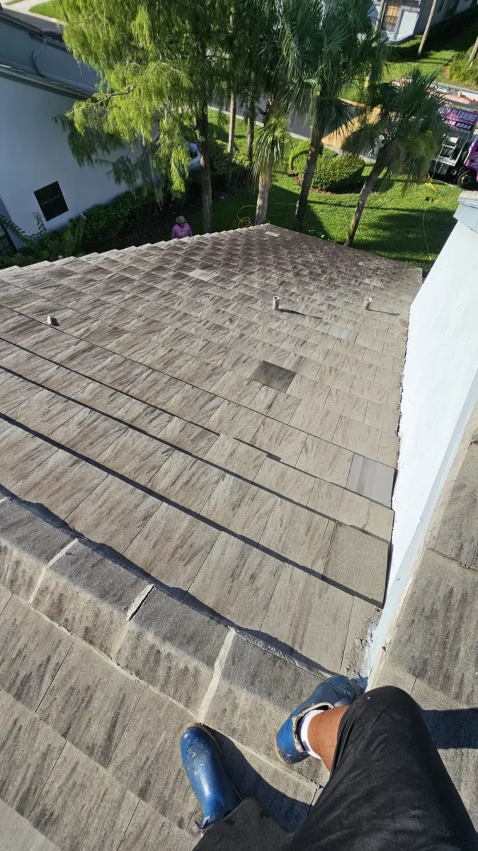 Let Us Restore Your Outdoor Living Space Person wearing blue shoes standing on a sloped, tiled roof with trees and green lawn nearby, likely during roofing or mainten